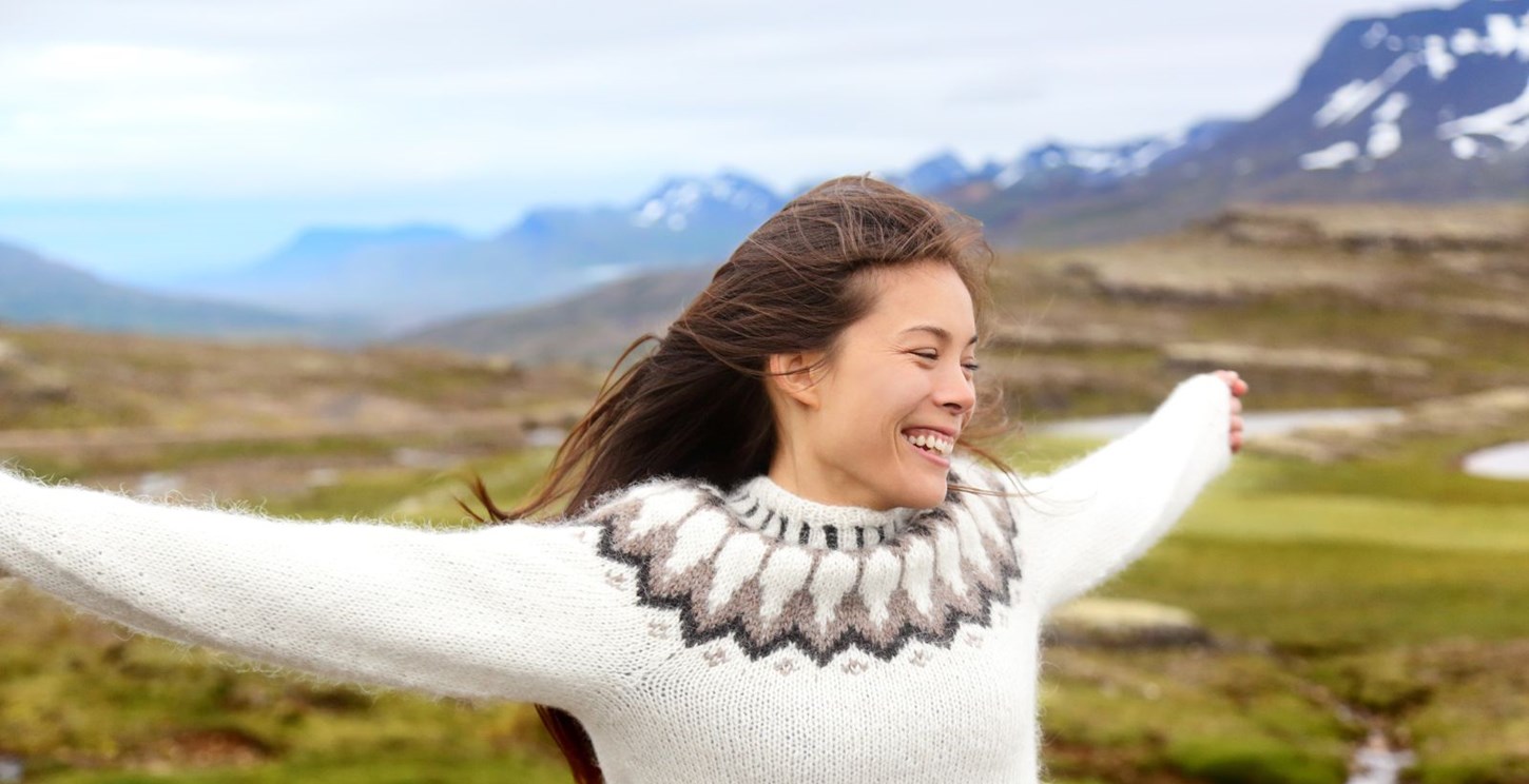 A woman wearing an Icelandic sweater, and smiling with her arms open in the Icelandic outdoors.