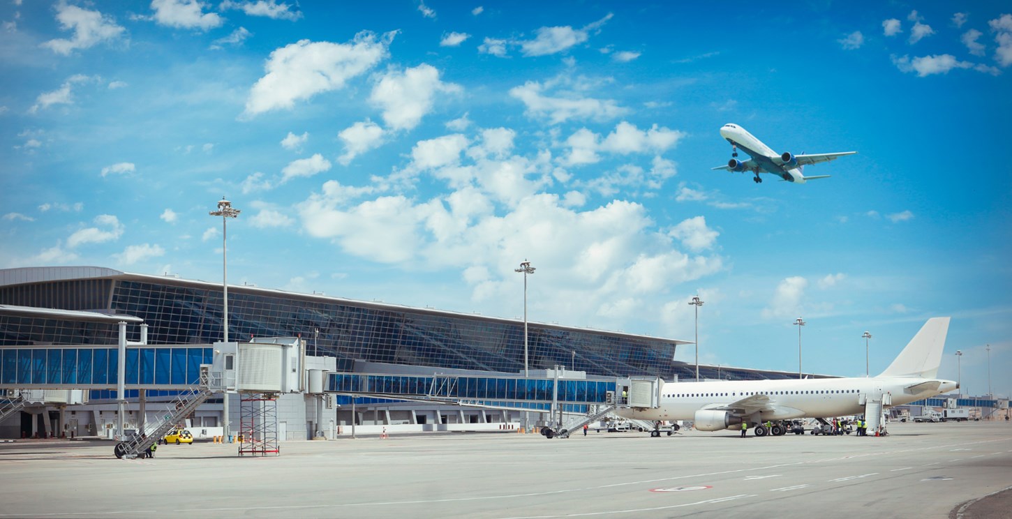 An airport with one aeroplane on the ground attached to a Passenger Boarding Bridge and another one in the sky.