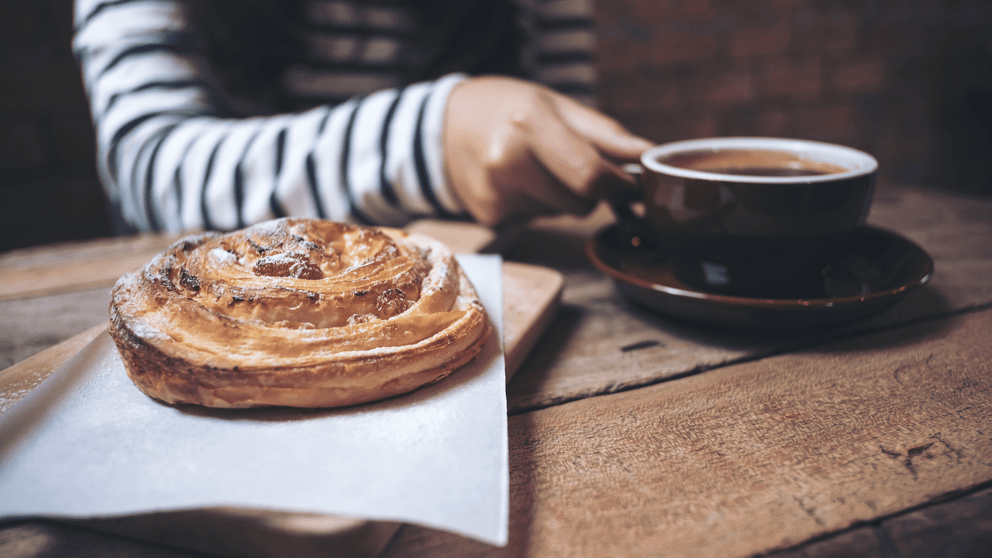 Woman holding a cup of coffee on a wooden table with a large pastry.