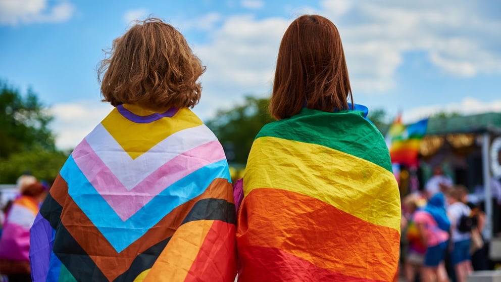 Two women wearing LQBTQ+ flags at a pride parade.