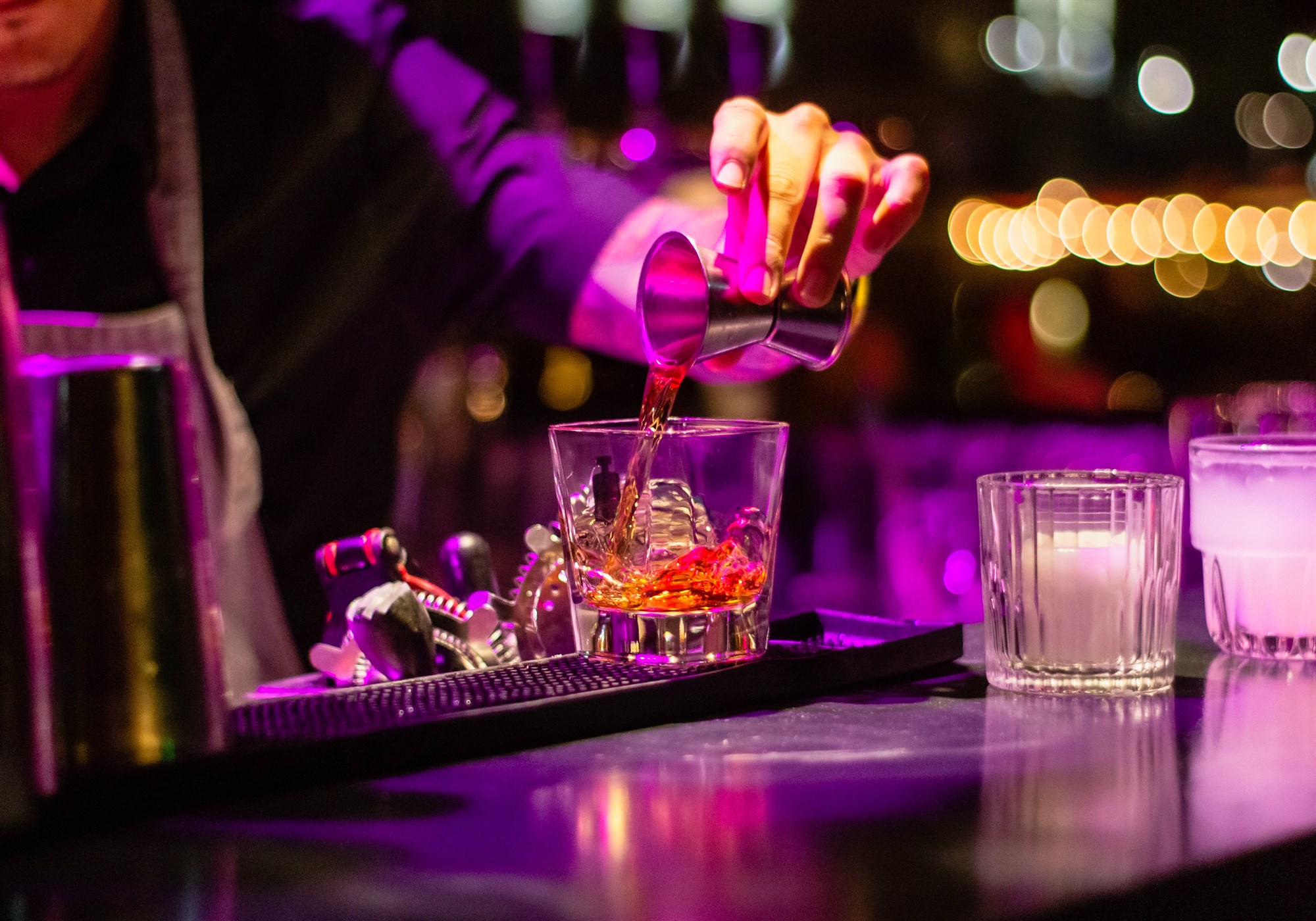 Bartender pouring a shot of spirit into a glass with ice on a bar top in purple light.