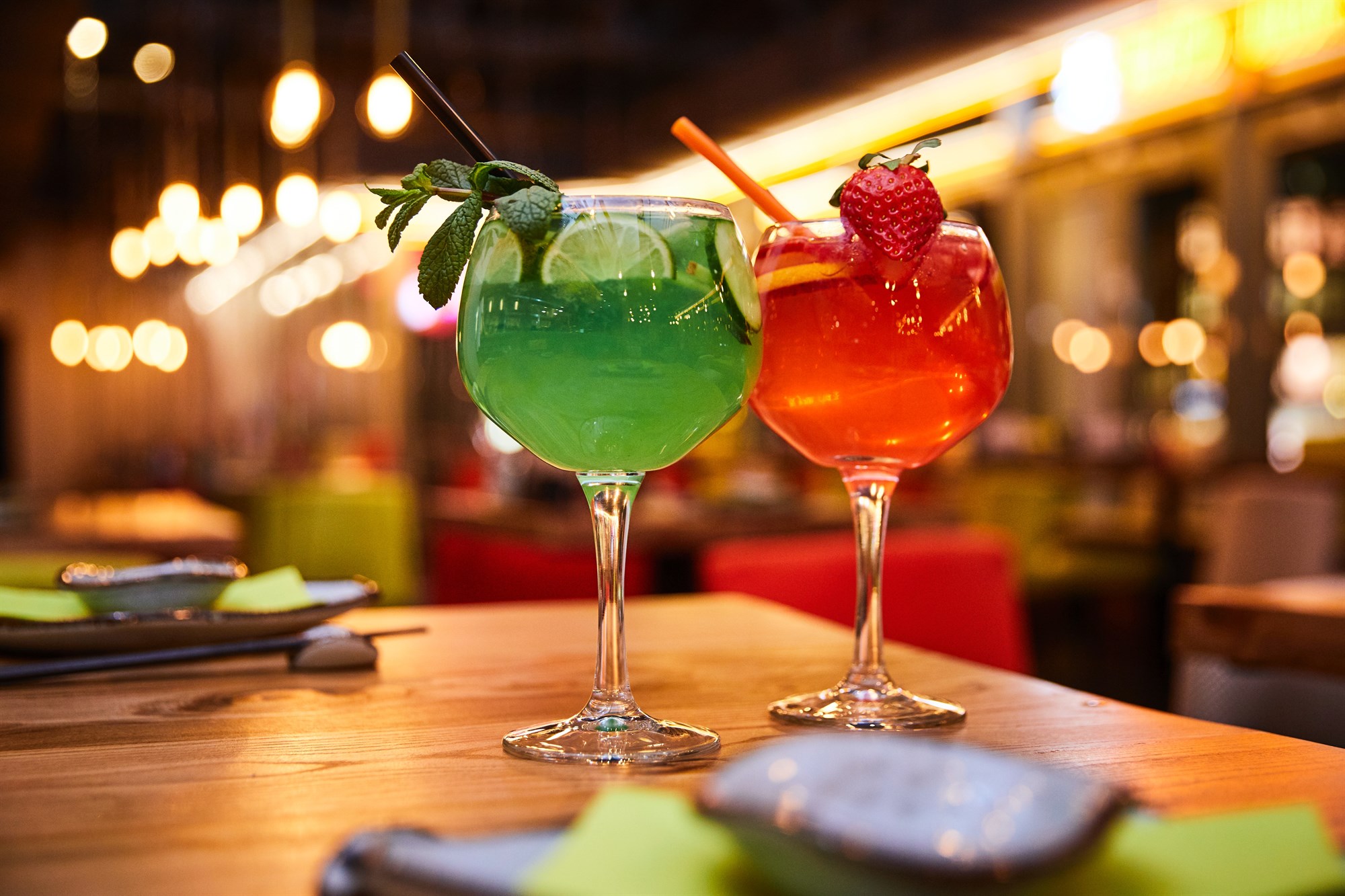 Two colourful cocktails on a table in a bar with fairy lights.