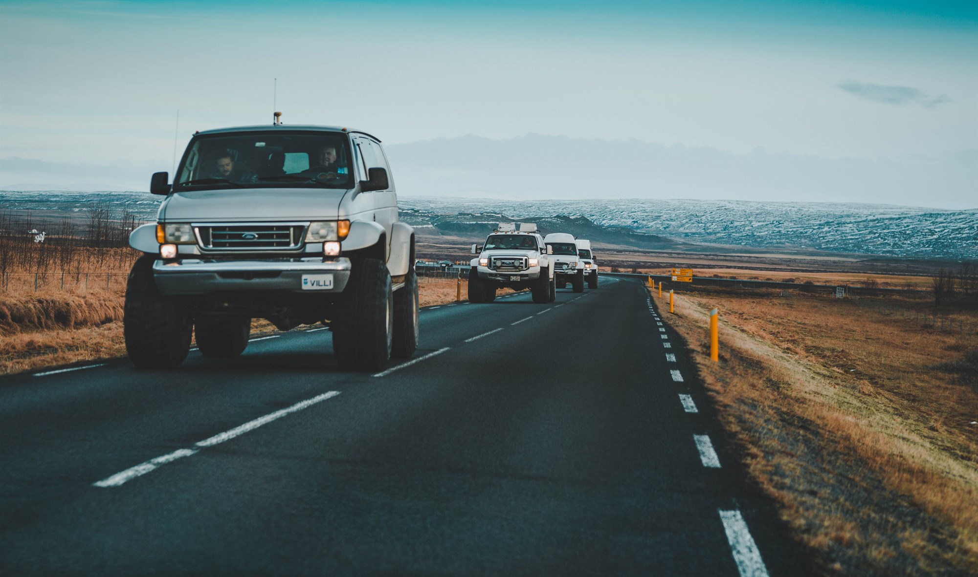 A convoy of large 4x4s on a road in Iceland with snow in the distance.