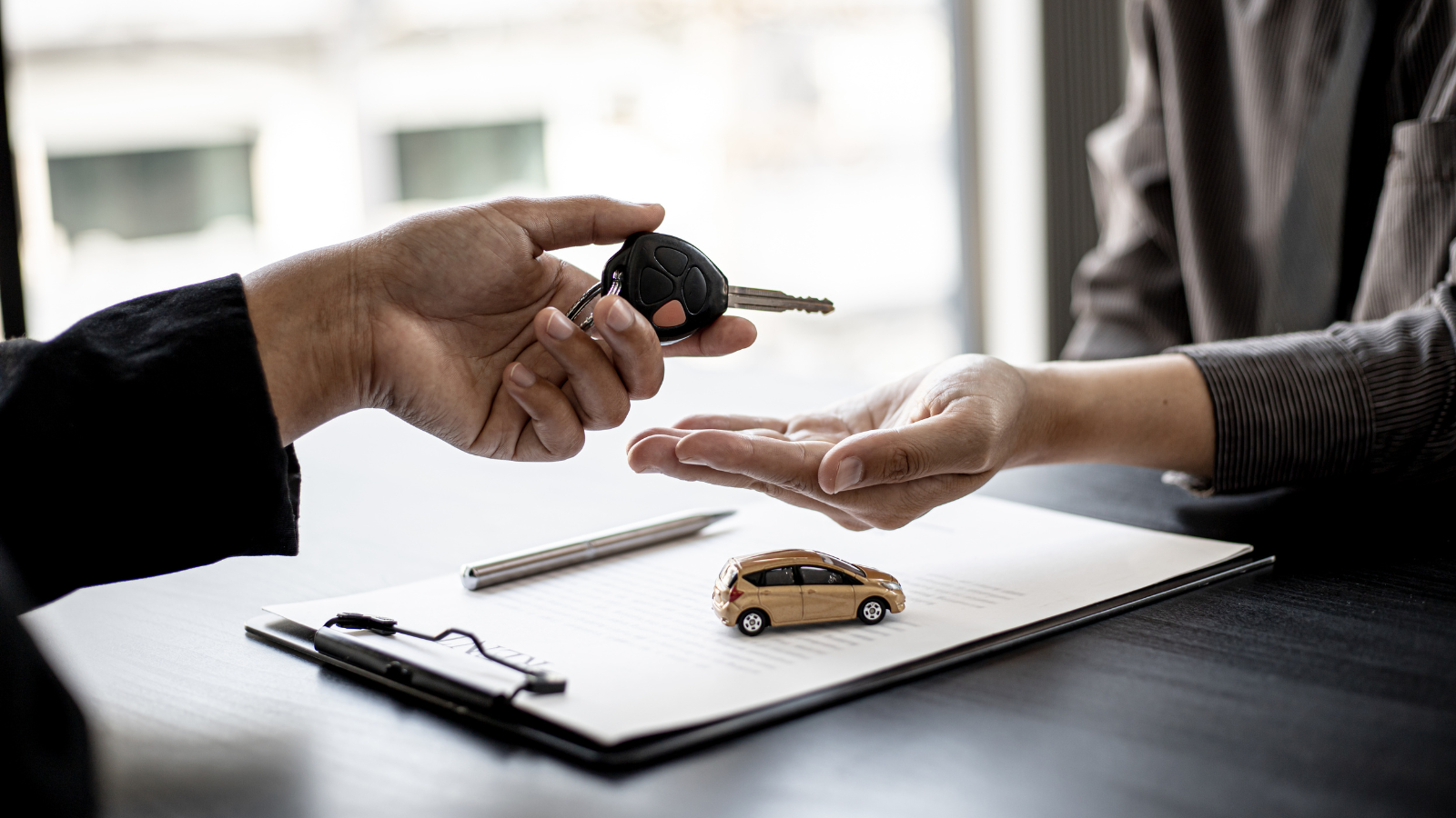 Two persons’ hands exchanging car keys over clipboard bearing a paper, a miniature car model, and some paper.