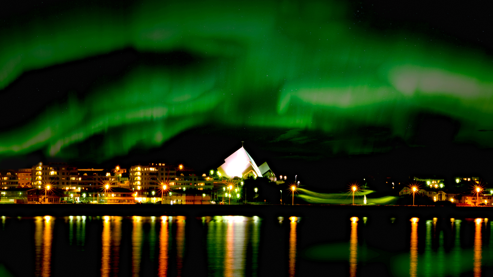 The Aurora Borealis over Reykjavík with the city lights reflected in the water at night.