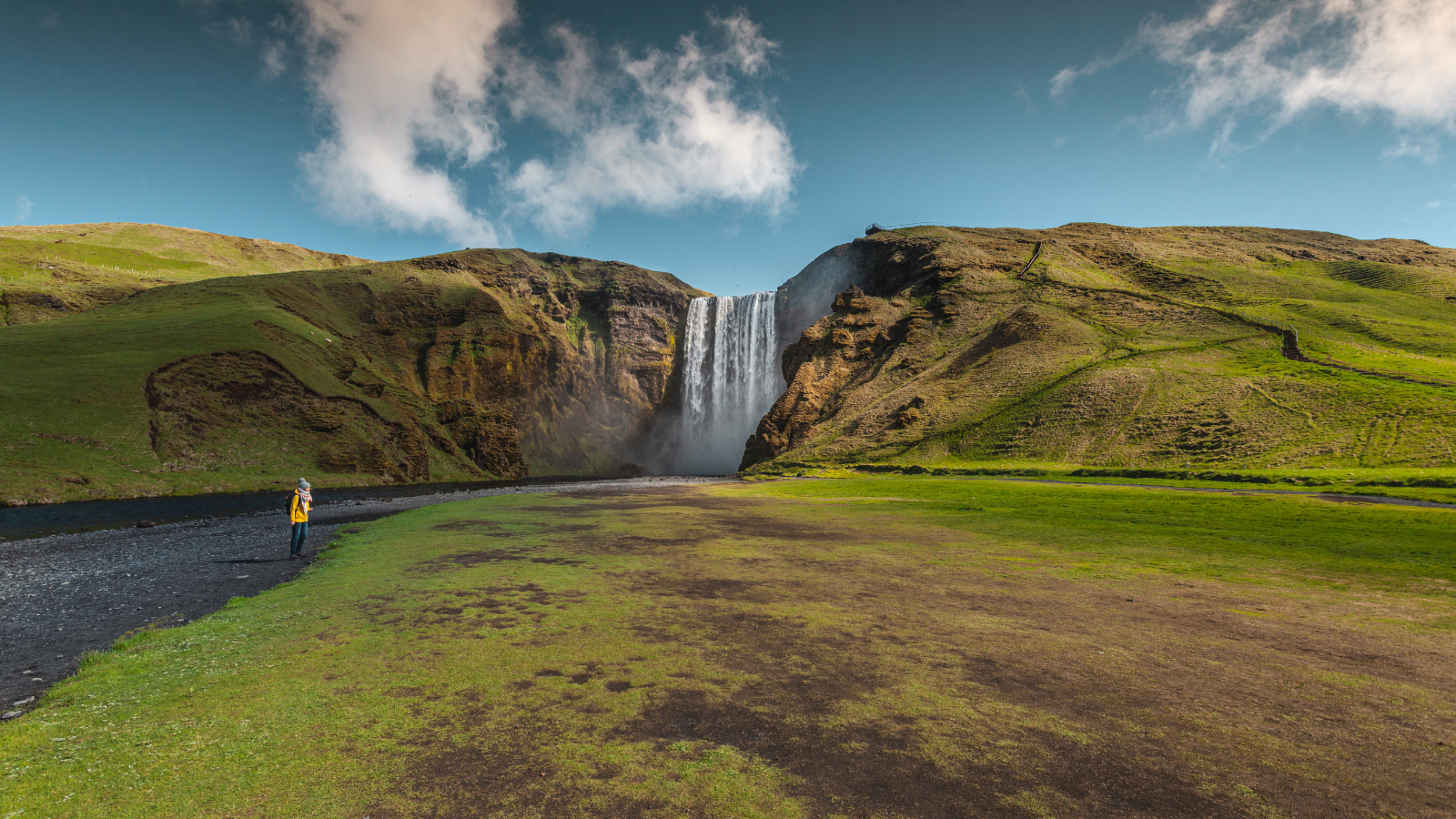 Grassy plain in front of Skógafoss with blue skies overhead.